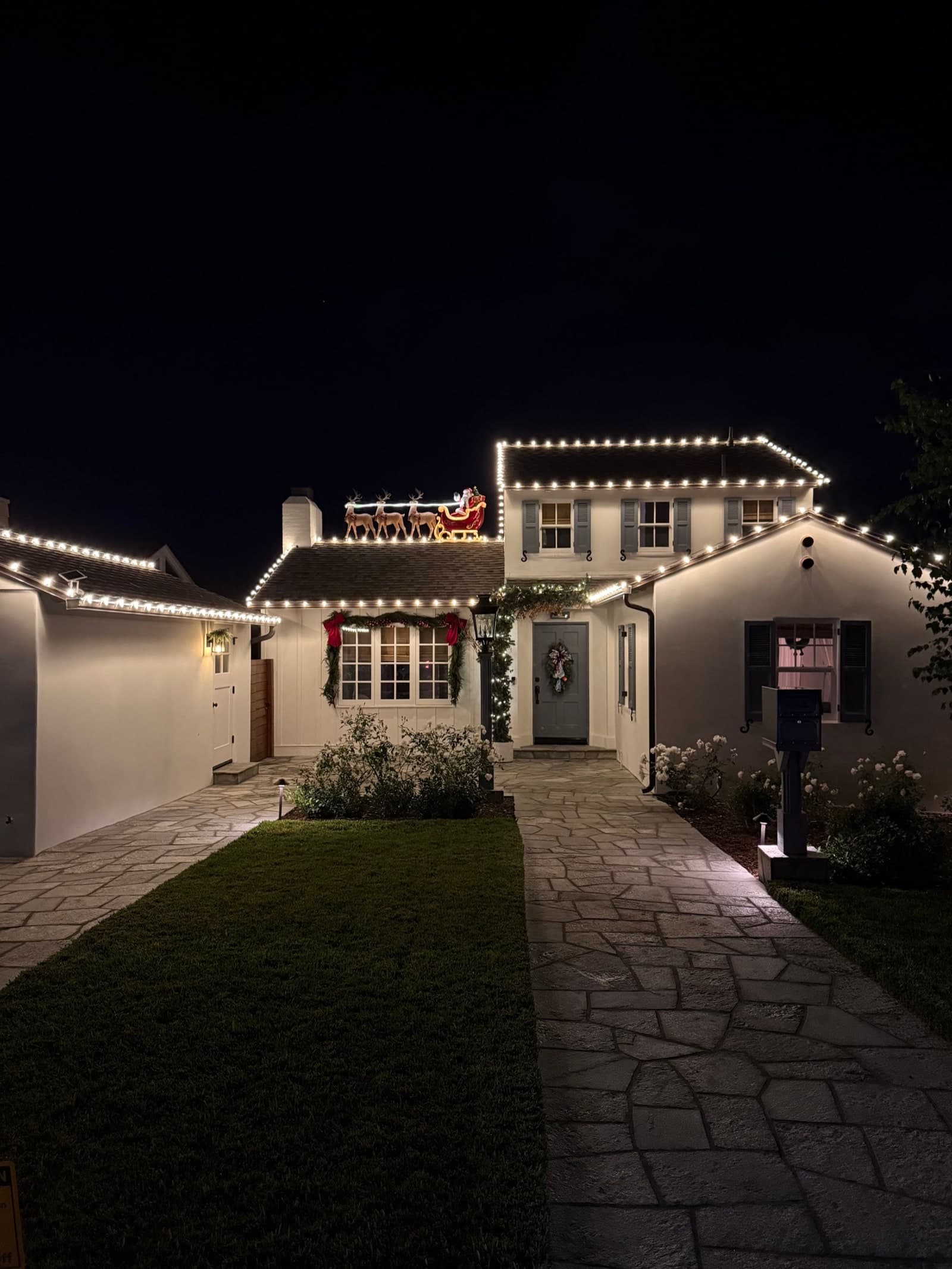 A La Jolla home at night with warm white C9 lights on every roofline, lit garland with red bows, and a Santa sleigh on the ridge